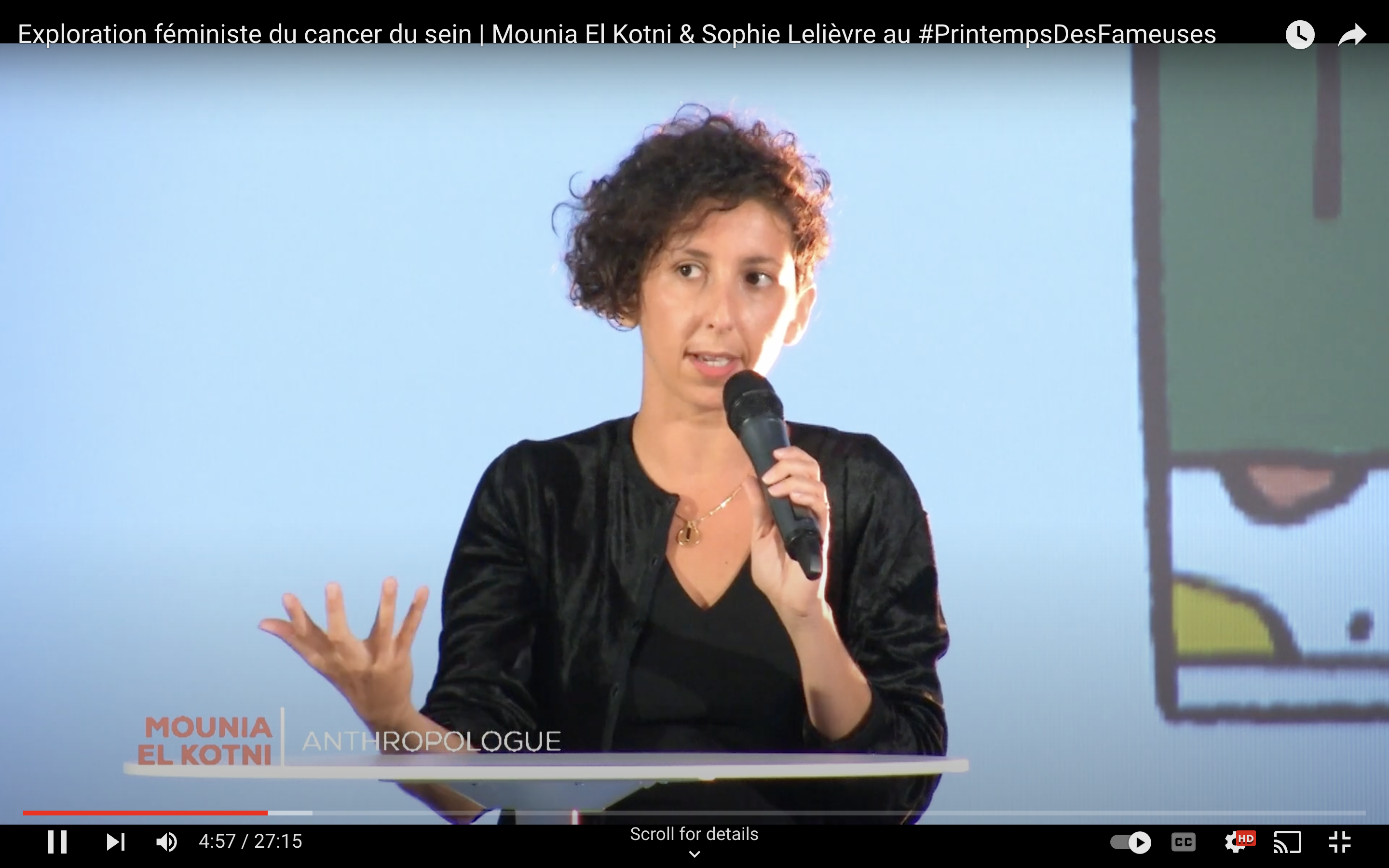 Femme brune aux cheveux courts habillée en noir, face à la caméra avec un micro dans la main gauche