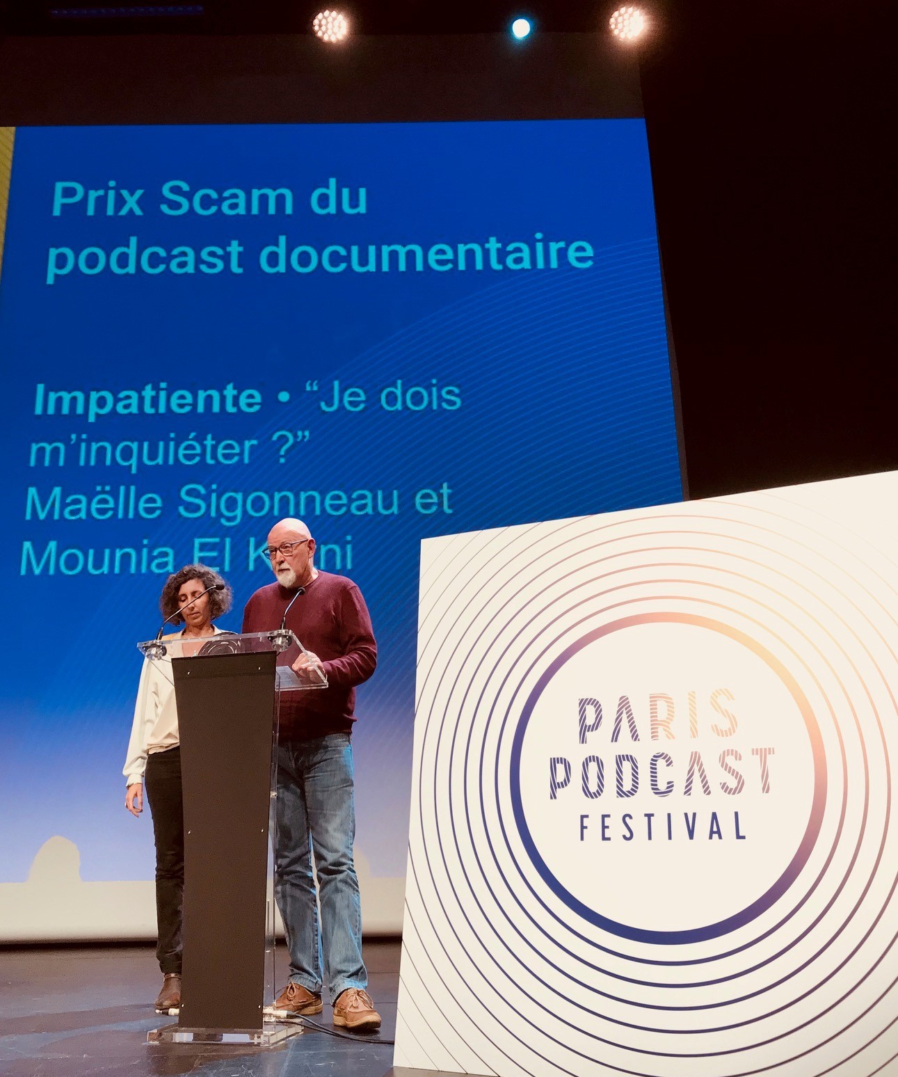 estrade du Paris Podcast Festival, logo au premier plan. Puis au second plan, Mounia El Kotni et Frédéric Sigonneau debout au micro, avec en fond un écran bleu où est écrit Prix Scam du podcast documentaire